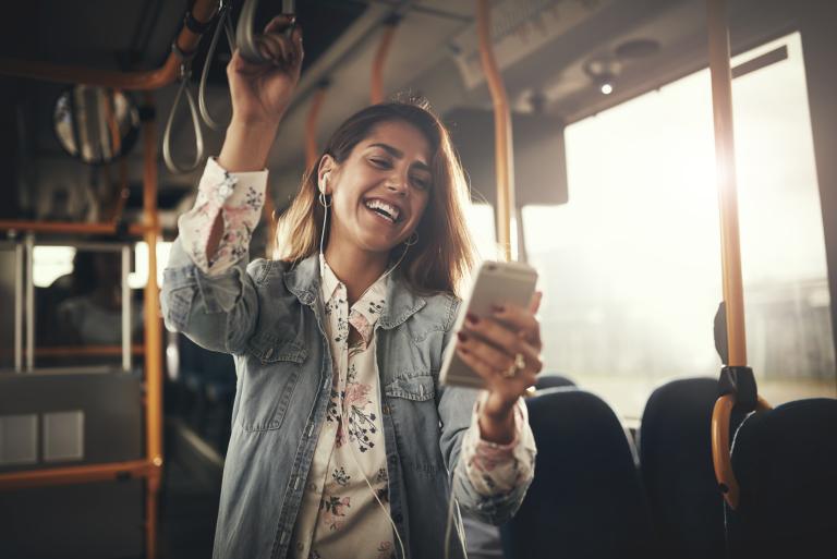 Young woman wearing earphones laughing at a text message on her cellphone while riding on a bus.